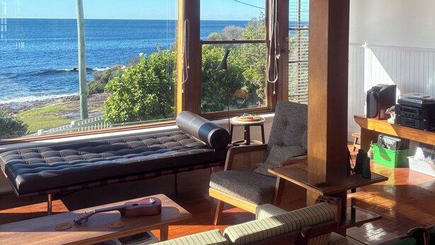 Une pièce dotée d’une grande fenêtre donnant sur l’océan, d’un canapé, d’un fauteuil, d’une petite table avec une guitare, et d’un meuble en bois contenant des haut-parleurs et des disques.