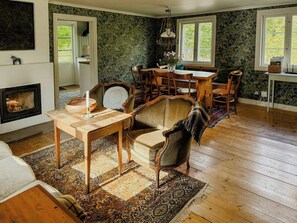 A cozy room with a fireplace, a dining table with chairs, and a large window. - Historic 19th century cottage in Blekinge (Kyrkhult)