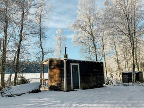 Une petite cabane en bois avec une cheminée, entourée d’arbres couverts de neige et sous un ciel dégagé.
