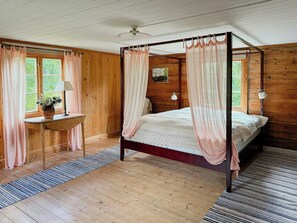 Un lit à baldaquin avec des rideaux légers, un plancher en bois, une petite table avec une lampe et une fenêtre également dotée de rideaux.