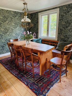 Un coin repas avec une table en bois, des chaises, un banc, un vase de fleurs, un tapis à motifs et une fenêtre donnant sur un paysage verdoyant.