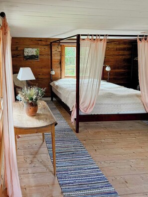 A wooden cabin bedroom with a four-poster bed, a table with a potted plant, and a window with a view of greenery. - Historic 19th century cottage in Blekinge (Kyrkhult)