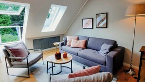 Ein modernes Wohnzimmer mit einem grauen Sofa, einem Sessel, einem kleinen runden Tisch und einer Stehlampe. Zwei Fenster mit Oberlichtern sind vorhanden, und im Hintergrund ist ein grauer Schrank zu sehen.