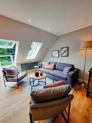 A modern living room with a grey sofa, a chair, a small round table, and a floor lamp. There are two windows with skylights, and a grey cabinet is visible in the background. - Romantic countryside in East Frisia (Friedeburg)