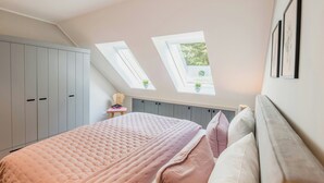 Ein Schlafzimmer mit schrägverdachter Decke, Dachfenstern, einem Bett mit rosa Bettdecke und grauen Stauraumeinheiten.