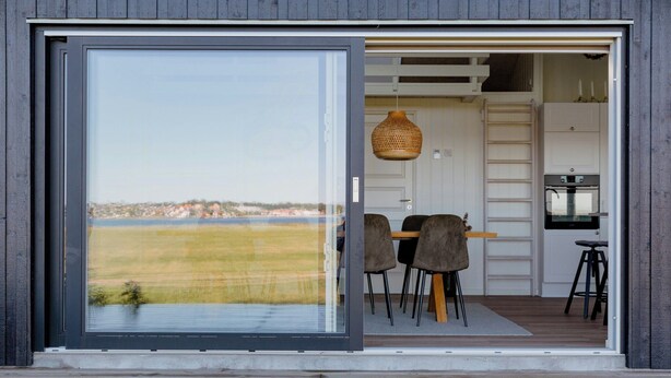 A modern interior with a large sliding glass door, a dining area with chairs and a table, a hanging light fixture, and a kitchen with a stove. - Fresh air on open terrace with view (Hejls)