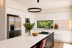 A modern kitchen with a built-in oven, a large window with a view of greenery, a central island with a bowl of lemons, and a hanging light fixture. - Luxury Athenian Riviera Villa with Private Pool (Vouliagmeni)