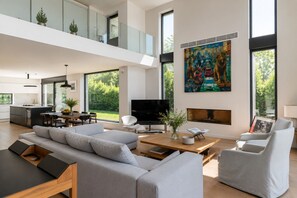 A modern living room with a sofa, chairs, a fireplace, a TV, and a dining area with a kitchen in the background. - Luxury Athenian Riviera Villa with Private Pool (Vouliagmeni)