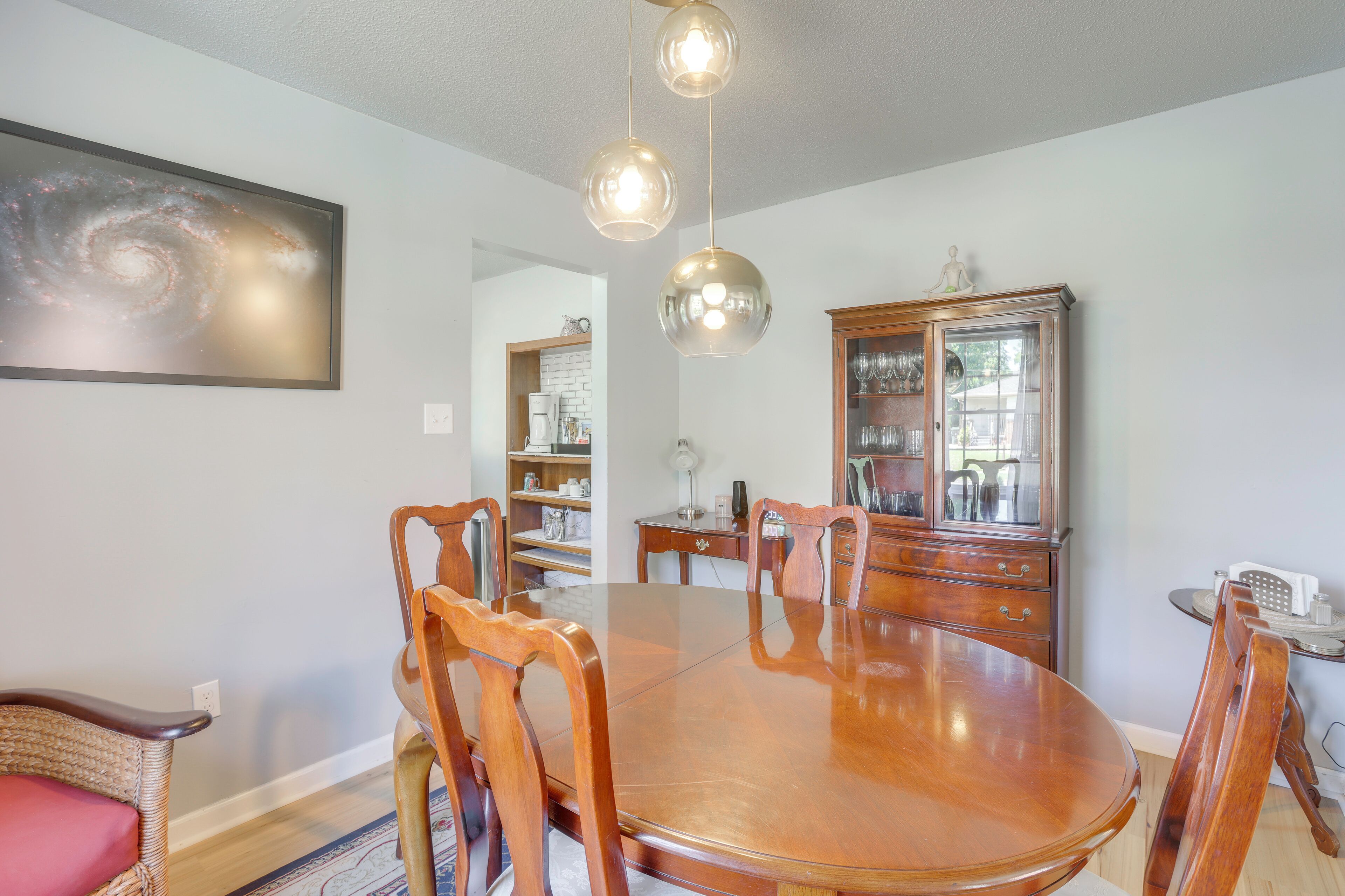 Een eetkamer met een ronde houten tafel, stoelen, een kroonluchter, een kast en een schilderij aan de muur.