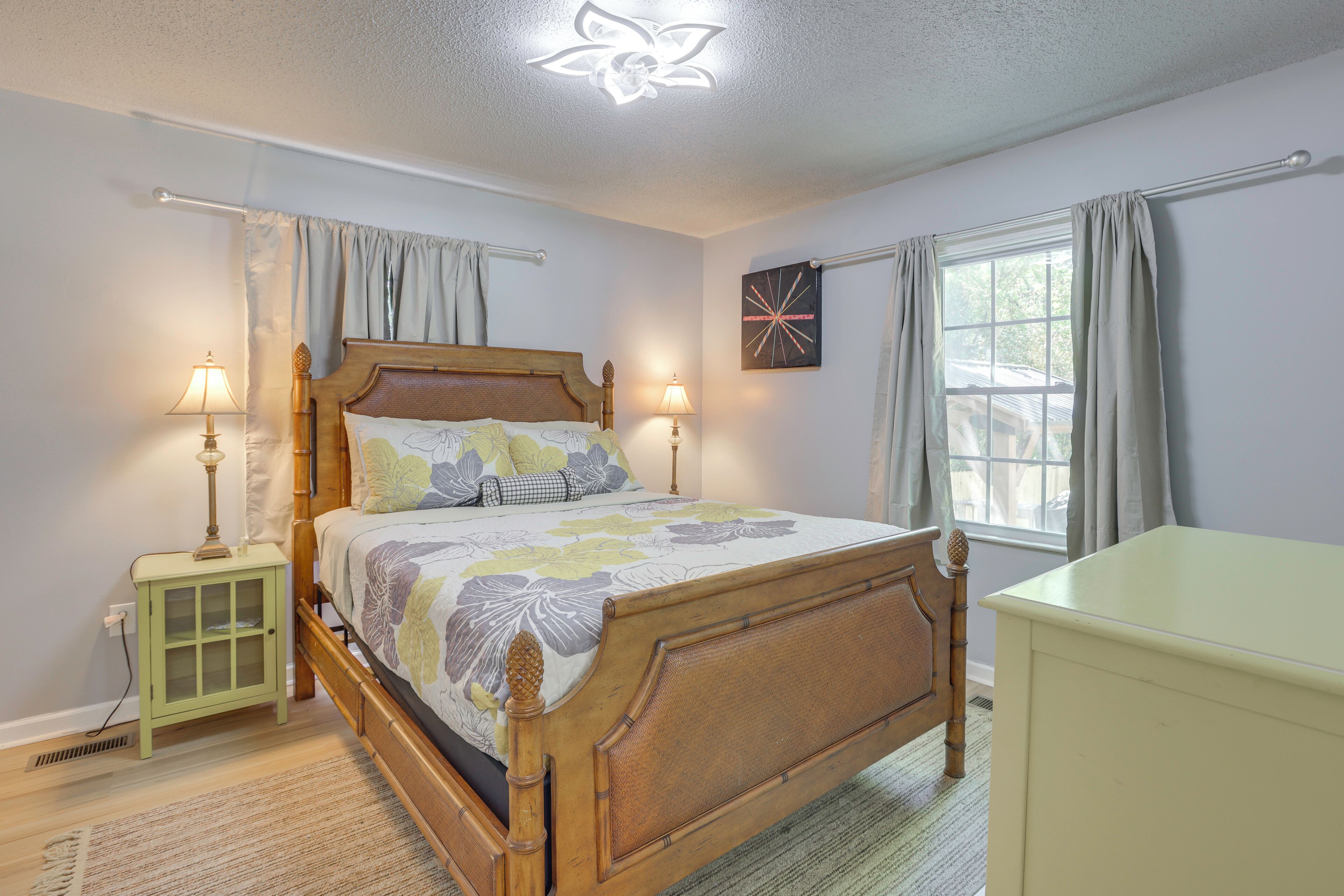 A bedroom with a wooden bed, bedside lamps, a window with curtains, and a wall-mounted artwork.