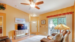 A living room with a ceiling fan, a television on a wooden cabinet, a sofa, and a window with a view of trees. - Resort-style Pools & Hot Tubs! Mtn-view Mesa Condo (Mesa)