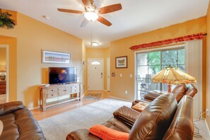 A living room with a ceiling fan, a television on a wooden cabinet, a sofa, and a window with a view of trees.