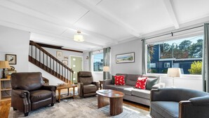 A spacious living room with a staircase, leather armchairs, a grey sofa with red pillows, and large windows offering a view of the outside. - Sand Dollar at Seaside Beach (Seaside)