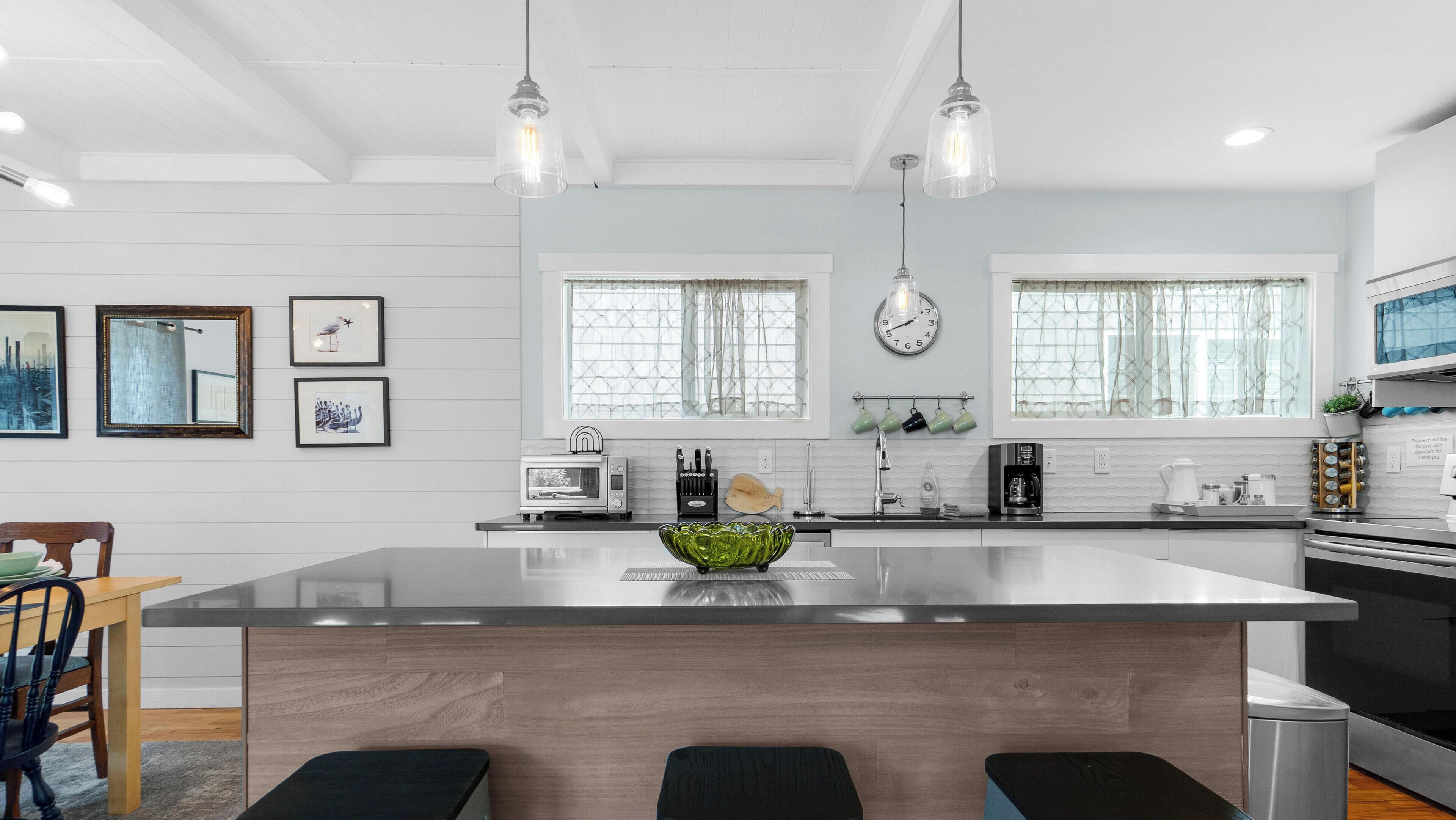 A modern kitchen with a large island, pendant lights, a clock, and framed artwork on the wall.