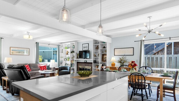 A modern kitchen with a central island, a dining area with a table and chairs, a living room with a sofa and chairs, and a large window with a view. - Sand Dollar at Seaside Beach (Seaside)