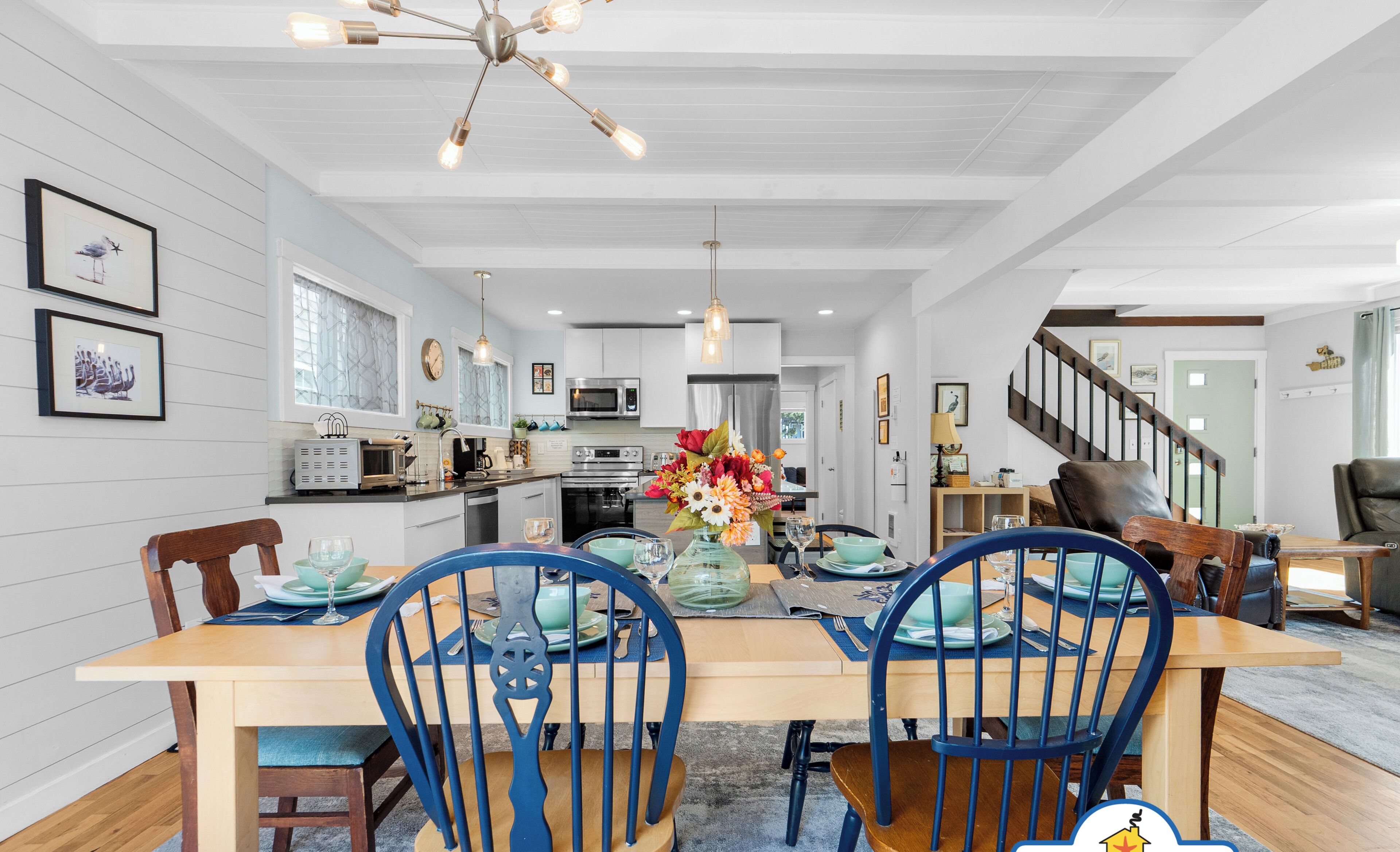 A dining area with a wooden table, blue chairs, a chandelier, and a kitchen in the background.