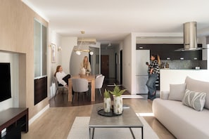 Ein modernes Wohnzimmer mit Sofa, Couchtisch und Fernseher. Es gibt einen Essbereich mit Tisch und Stühlen. Im Hintergrund ist eine Küche mit Schränken und einem Herd zu sehen.