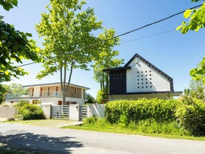 Uma casa moderna de dois andares com um portão na entrada, cercada por vegetação exuberante e um céu azul límpido.