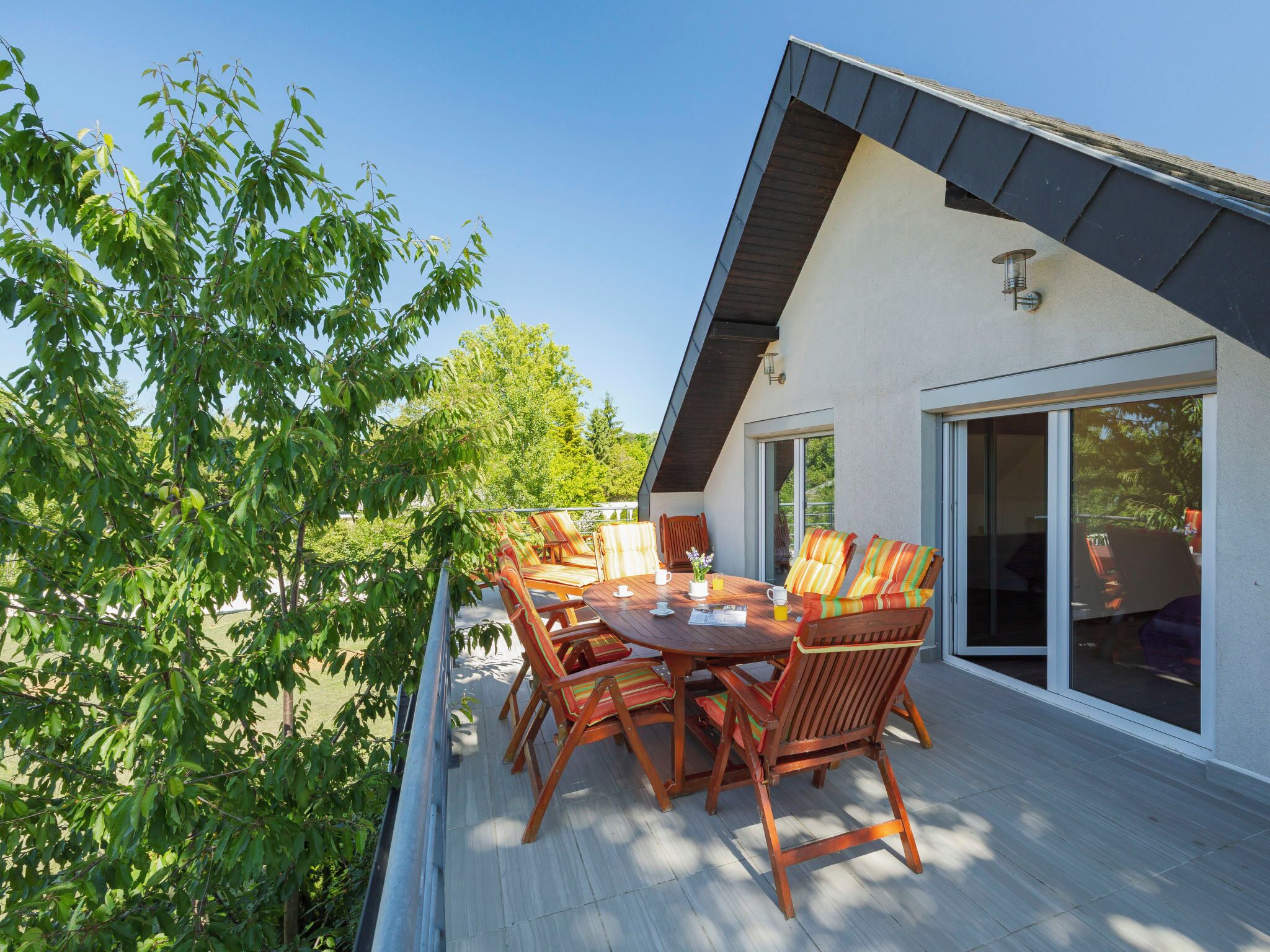A modern outdoor dining area with a wooden table and chairs, a glass door leading inside, and a view of the surrounding greenery.