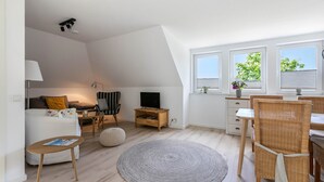 A modern living room with a sofa, TV, wooden side table, and a dining area with wicker chairs. - NEW - Artist’s Retreat Halebüll with Sea View, Shared Terrace and Wi-Fi (Husum)