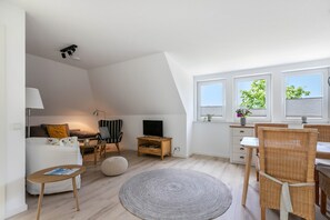 A modern living room with a sofa, TV, wooden side table, and a dining area with wicker chairs.