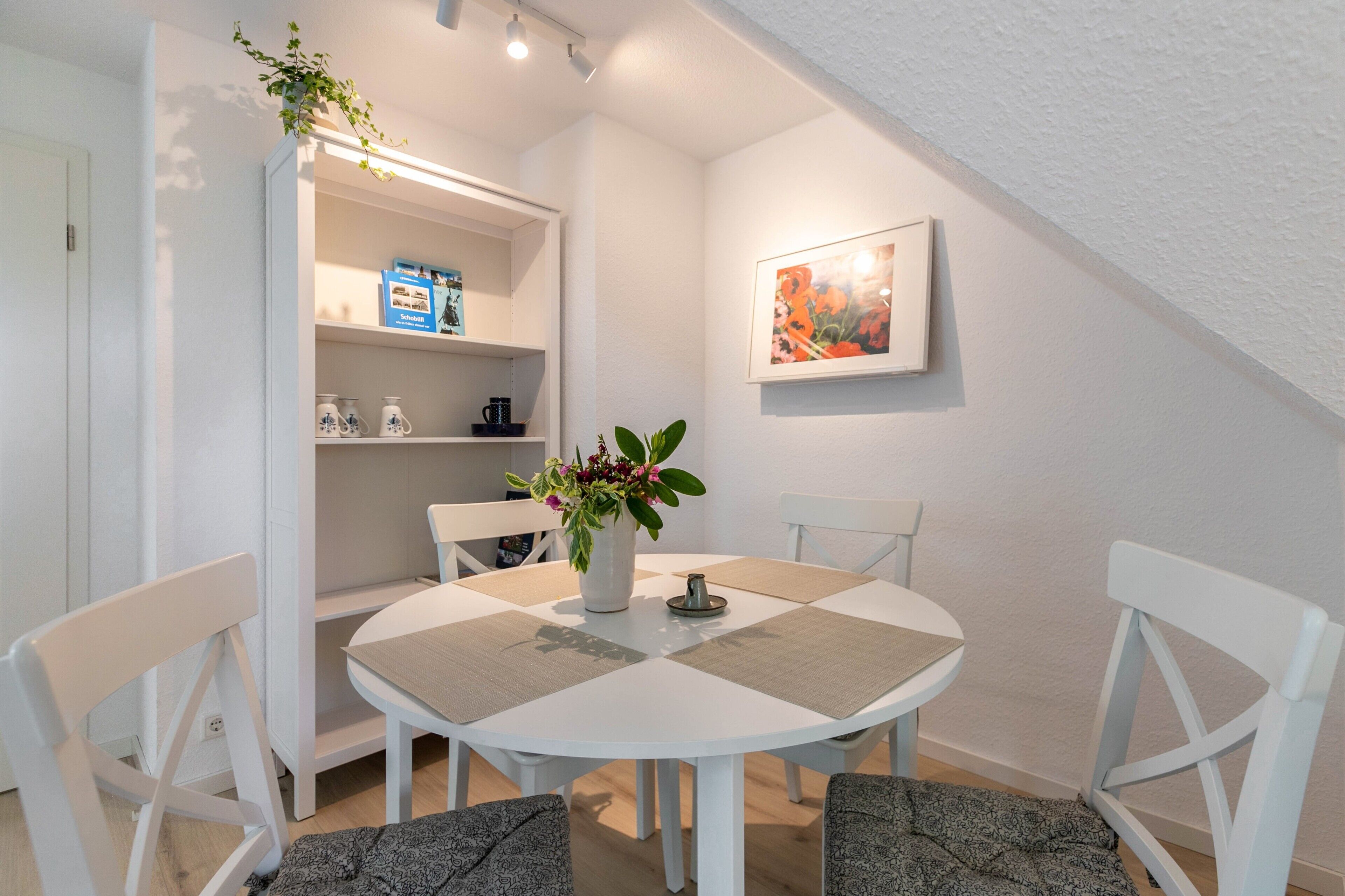 A round dining table with white chairs, a potted plant, and a shelf with kitchenware.