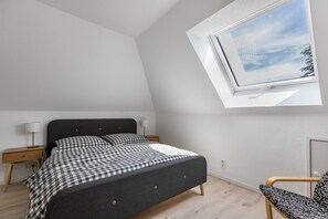 A bedroom with a bed, a nightstand, a chair, a window with a view of the sky, and a door.