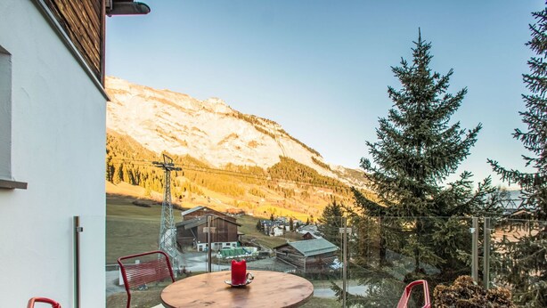 Een balkon met een ronde tafel en rode stoelen, met uitzicht op een bergachtig landschap met een houten chalet en een hoge metalen constructie.