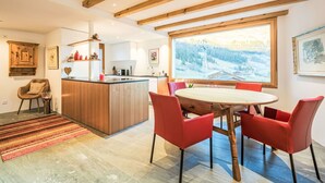 Een moderne keuken met houten balken, een eethoek met rode stoelen en een groot raam met uitzicht op sneeuw.