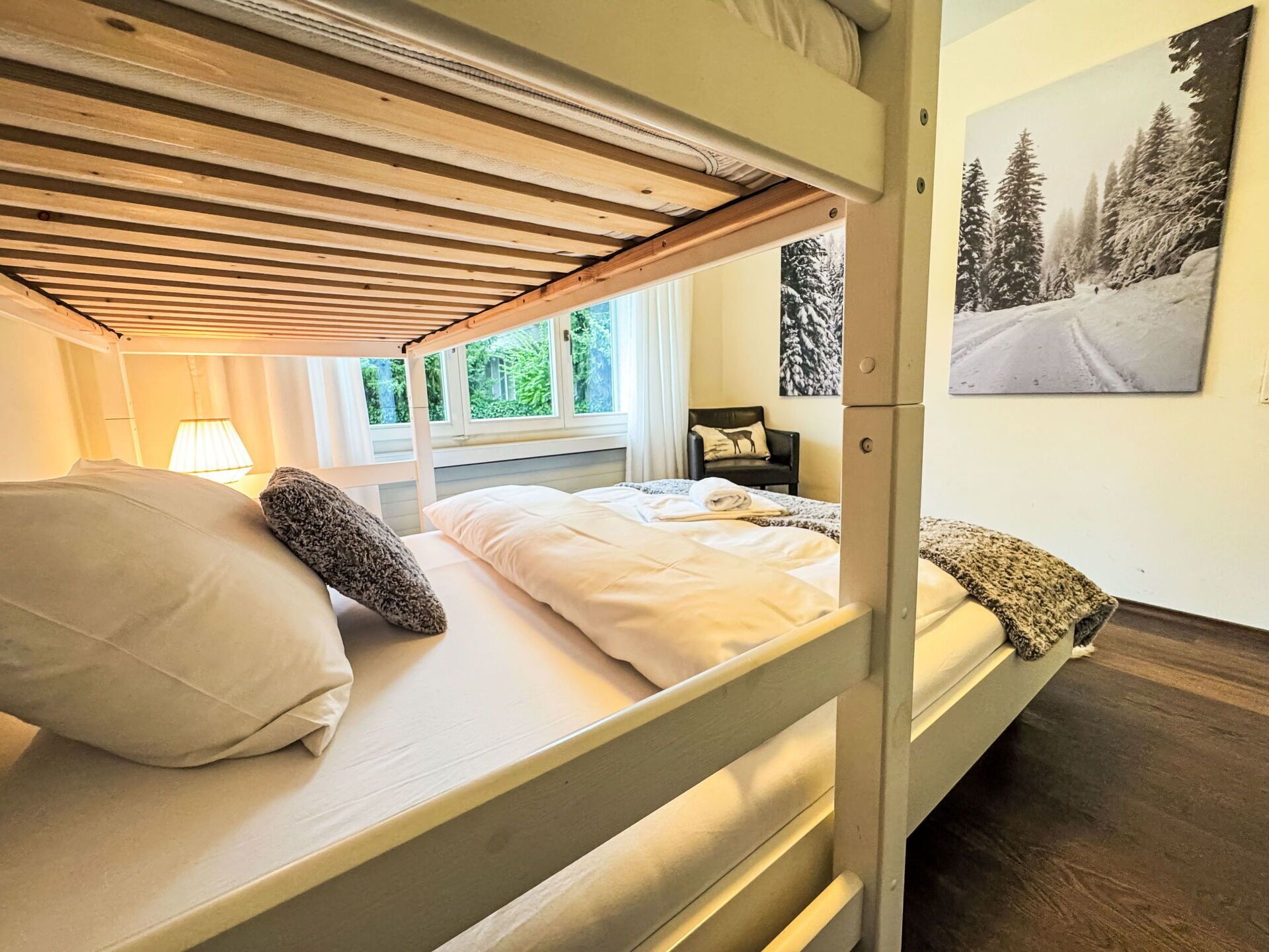 A bunk bed with a wooden ceiling, a window with a view of greenery, a chair with a cushion, and a painting of a snowy landscape.