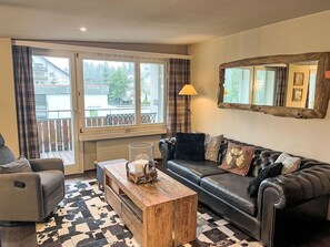 A living room with a sofa, a wooden coffee table, a lamp, and a large window with curtains.