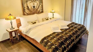 Een net opgemaakt bed met witte lakens, een gebreide deken met een patroon en twee kussens met dierenprints. Daarboven hangt aan de muur een lijst met een afbeelding van een wolf, en ernaast een klein tafeltje met een lamp.