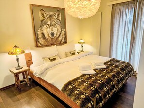 Ein ordentlich bezogenes Bett mit weißen Bettwäsche, einer gemusterten Tagesdecke und zwei Kissen mit Tiermotiven. An der Wand über dem Bett hängt ein gerahmtes Bild von einem Wolf, daneben steht ein kleiner Tisch mit einer Lampe.