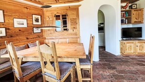 A wooden dining area with a table and chairs, a kitchen in the background, and a rustic ceiling.