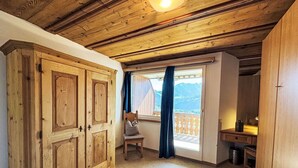 A room with wooden ceilings, a large wardrobe, a chair, a desk, and a view of mountains through a window.