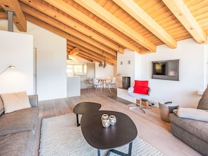 Ein modernes Wohnzimmer mit Holzbalkendecke, einem schwarzen Couchtisch mit Kerzen, einem grauen Sofa und einem roten Akzentstuhl.