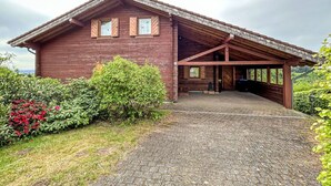 Une cabane en bois dotée d’un porche couvert, d’une allée en gravier et entourée de verdure.