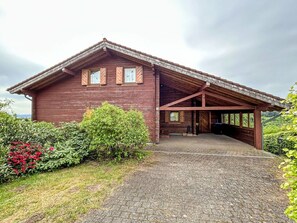 Eine Holzhütte mit verdeckter Veranda, einem Schotterweg und umliegender Bepflanzung.