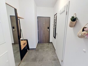 Un couloir avec une porte en bois, un miroir, une plante fixée au mur et un petit panier rempli de fleurs.