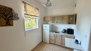 A compact kitchen with a refrigerator, a sink, a kettle, and a coffee maker. There is a window with a view of trees and a wall-mounted artwork of a tree. - MeThea SeaView Apartments by Greek Pride (Kassandra)