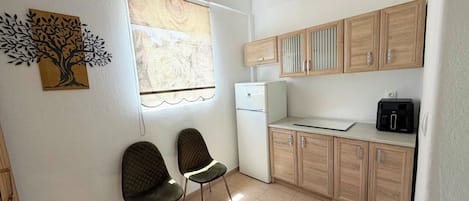 A compact kitchen with two chairs, a refrigerator, wooden cabinets, and a wall-mounted light fixture.