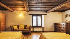Ein Zimmer mit Holzbalken, einem Korbssessel, zwei Betten mit gelber Bettwäsche, einem Schminktisch und einem Fenster mit Blick.