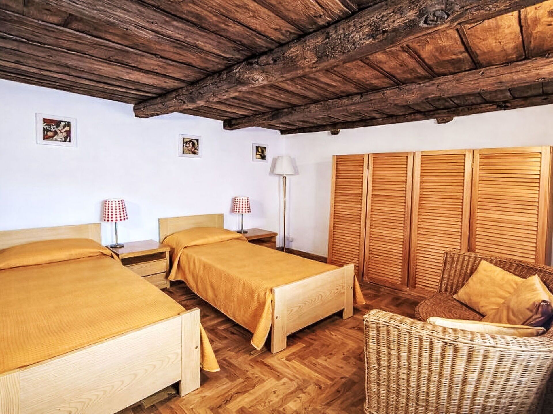 A room with two beds, a wicker chair, wooden wardrobe, and a wooden ceiling.