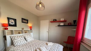 A bedroom with a bed, bedside lamps, a red chair, a door, and a shelf with boxes. - Les appartements de katia  (Charnay-lès-Mâcon)