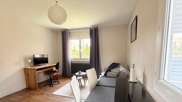 A compact room with a wooden desk, a TV, a dining table with chairs, a radiator, and a window with curtains. - Les appartements de katia  (Charnay-lès-Mâcon)