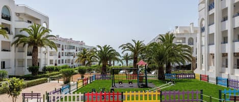 Une cour intérieure agrémentée de palmiers, une aire de jeux et un bâtiment blanc doté de balcons.