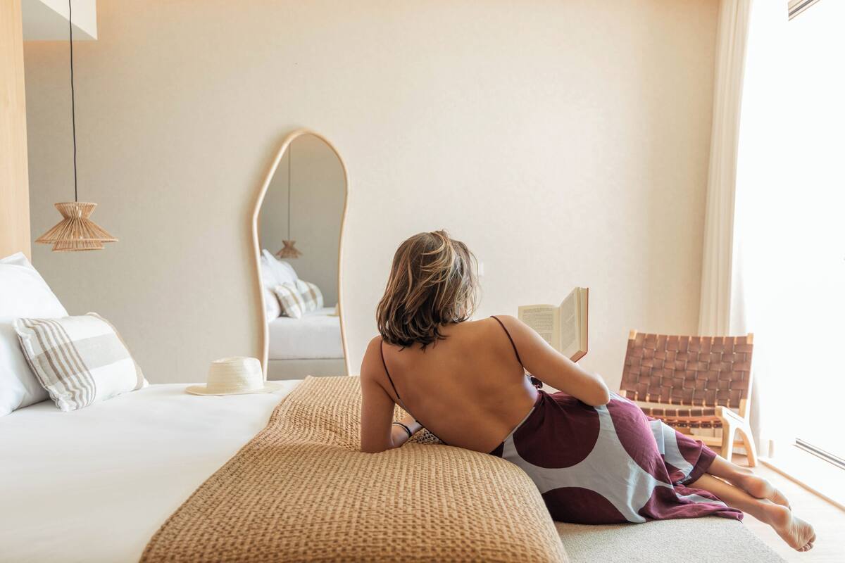 A person sitting on a bed in a room with a large mirror, a wicker chair, and a bedside table with a lamp.