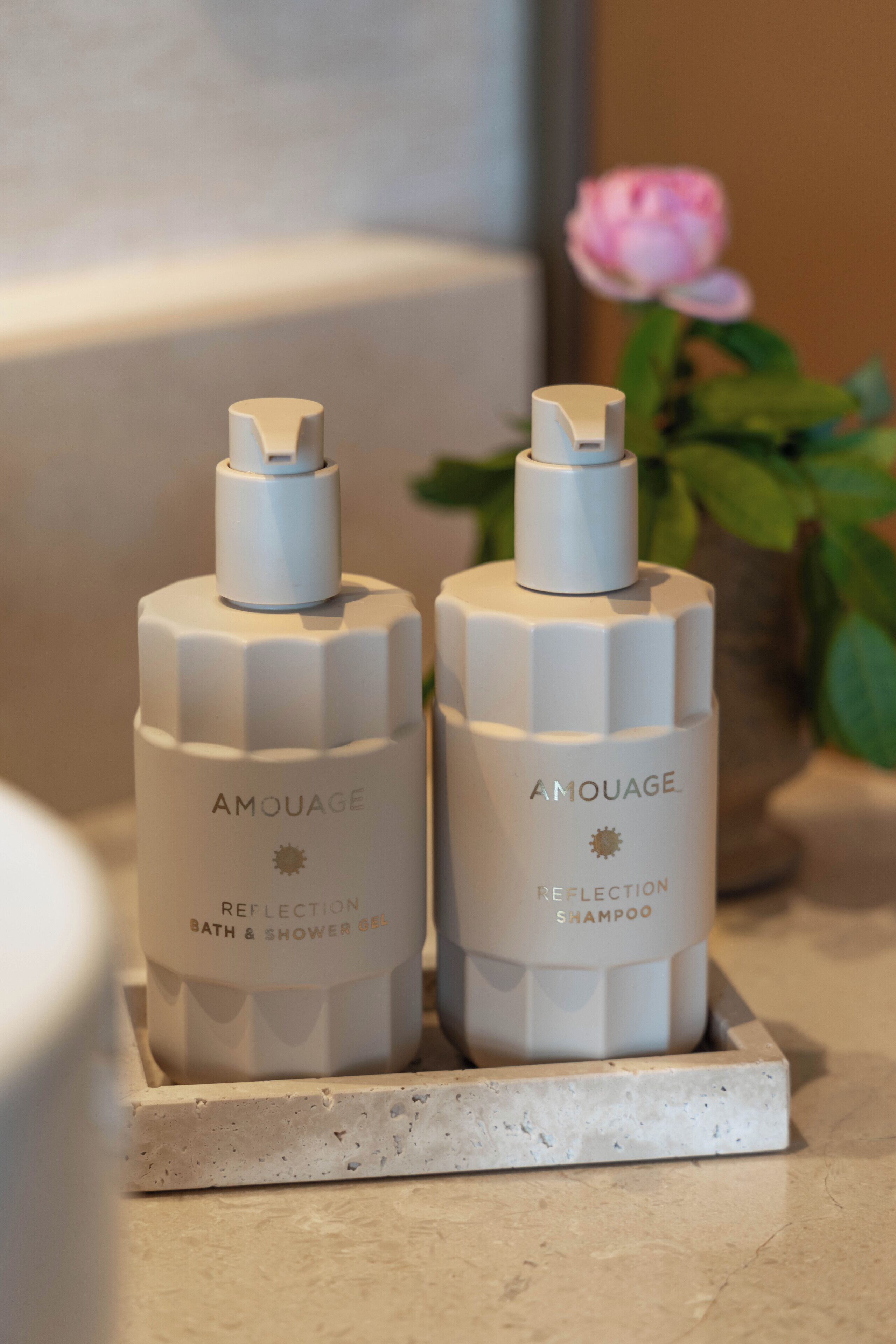 Two Amouage Reflection Bath & Shower Gel bottles and one Amouage Reflection Shampoo bottle on a marble surface with a potted plant in the background.