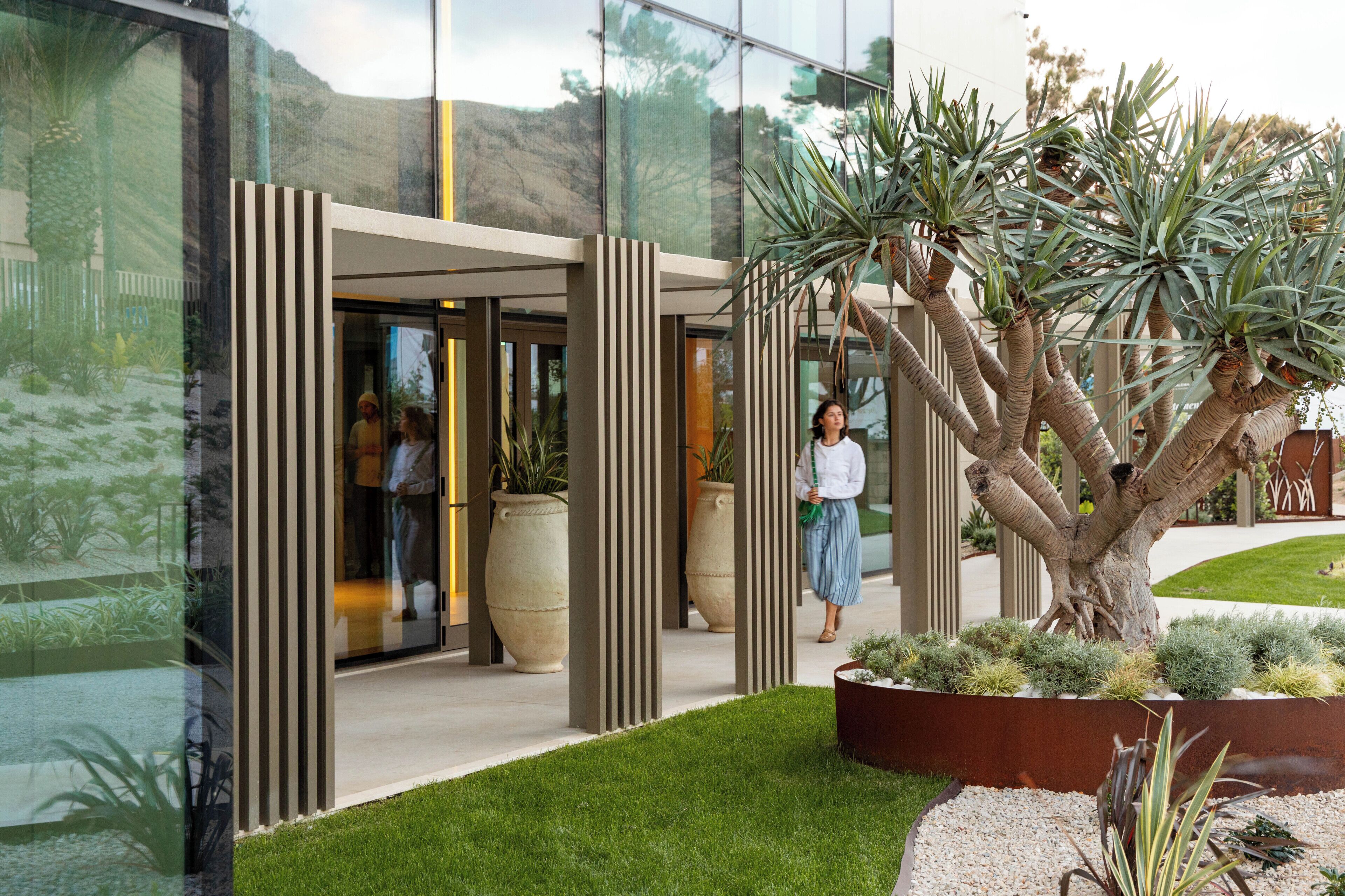 A modern building entrance with vertical columns, a glass facade, and a well-maintained lawn with various plants.