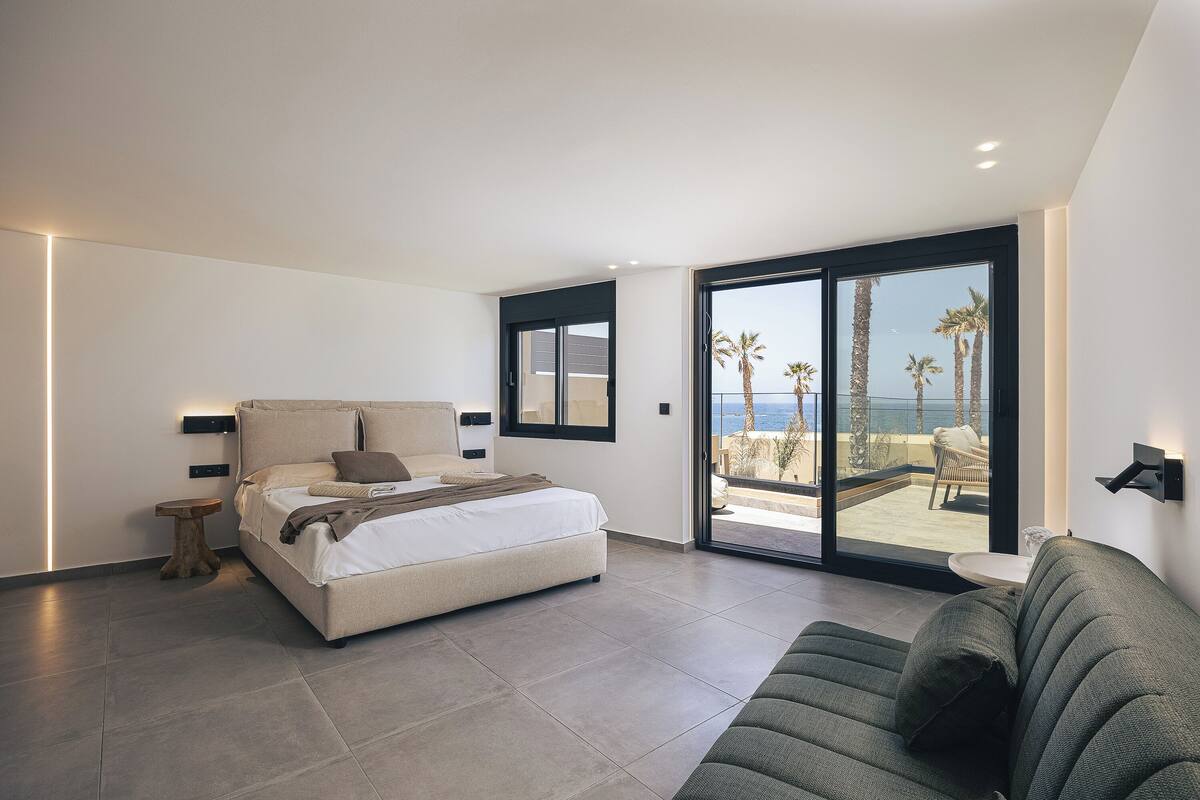 A modern bedroom with a bed, a green sofa, a small wooden side table, and a balcony with a view of palm trees and the sea.